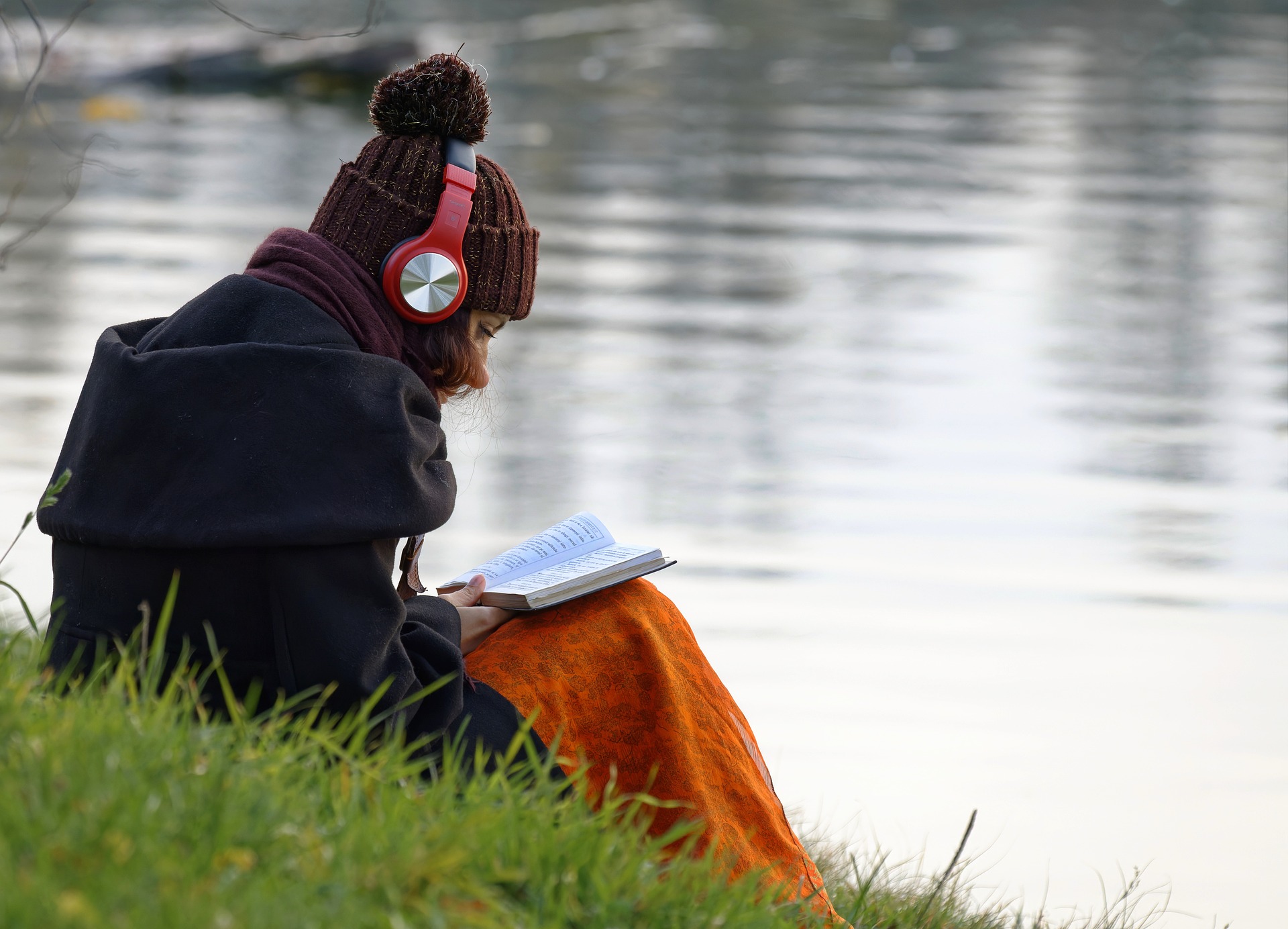 Eine Frau sitzt mit Kopfhörern und einem Buch an einem See.